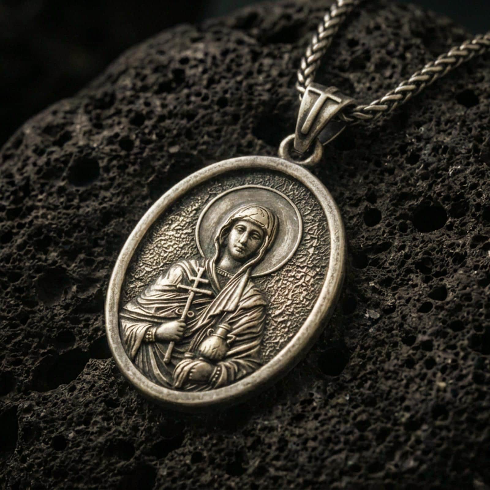 Bronze Saint Anastasia pendant with haloed saint figure, cross detail, and oval devotional portrait displayed on dark volcanic rock in a close-up luxury jewelry composition