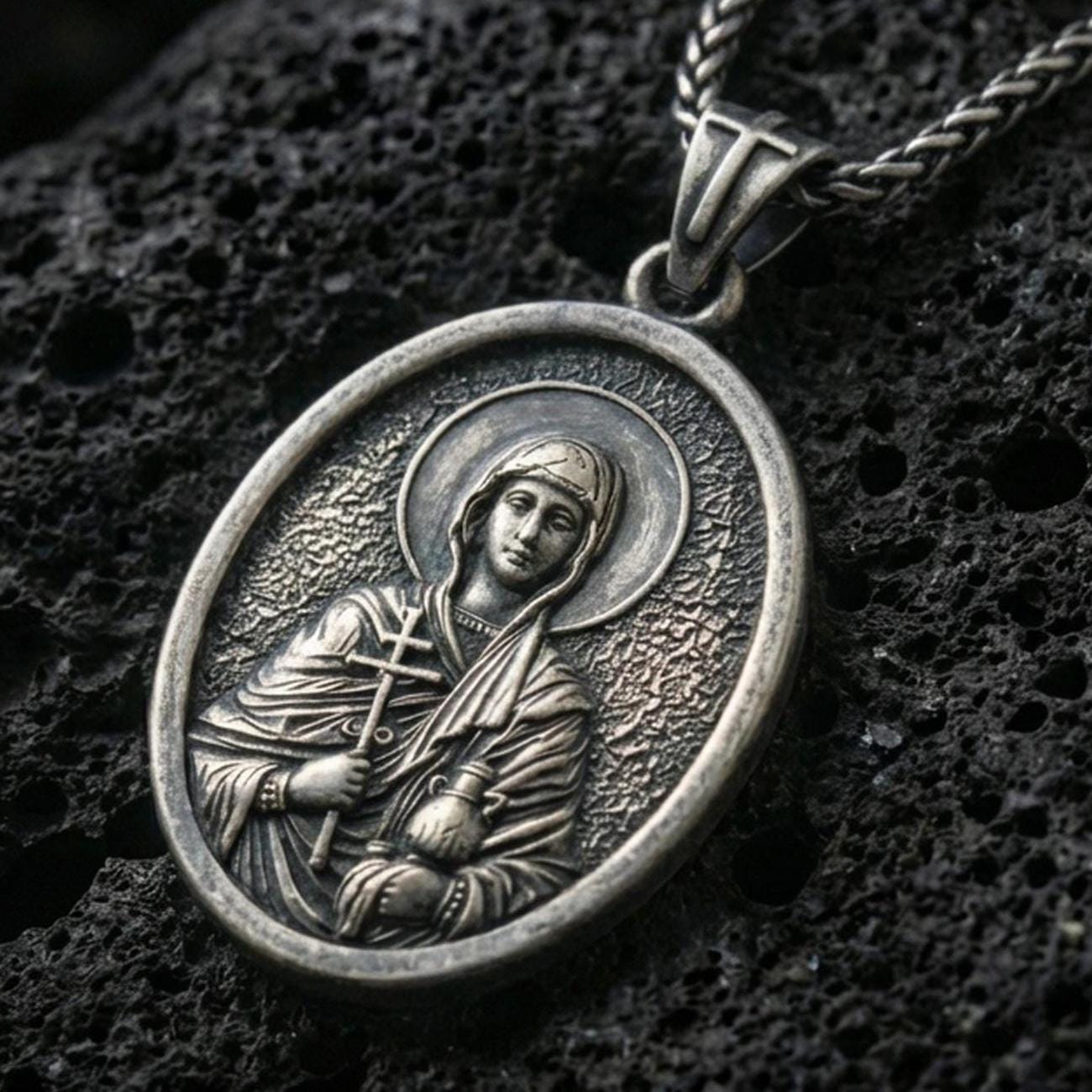 Oxidized Saint Anastasia pendant with haloed saint figure, cross detail, and oval devotional portrait displayed on dark volcanic rock in a close-up luxury jewelry composition