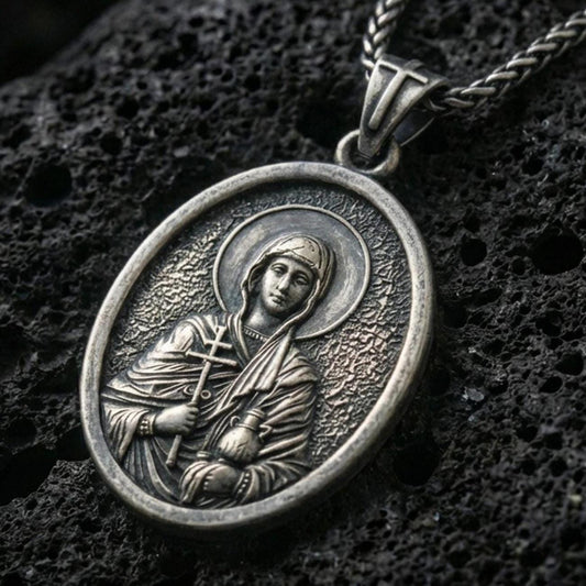 Oxidized Saint Anastasia pendant with haloed saint figure, cross detail, and oval devotional portrait displayed on dark volcanic rock in a close-up luxury jewelry composition