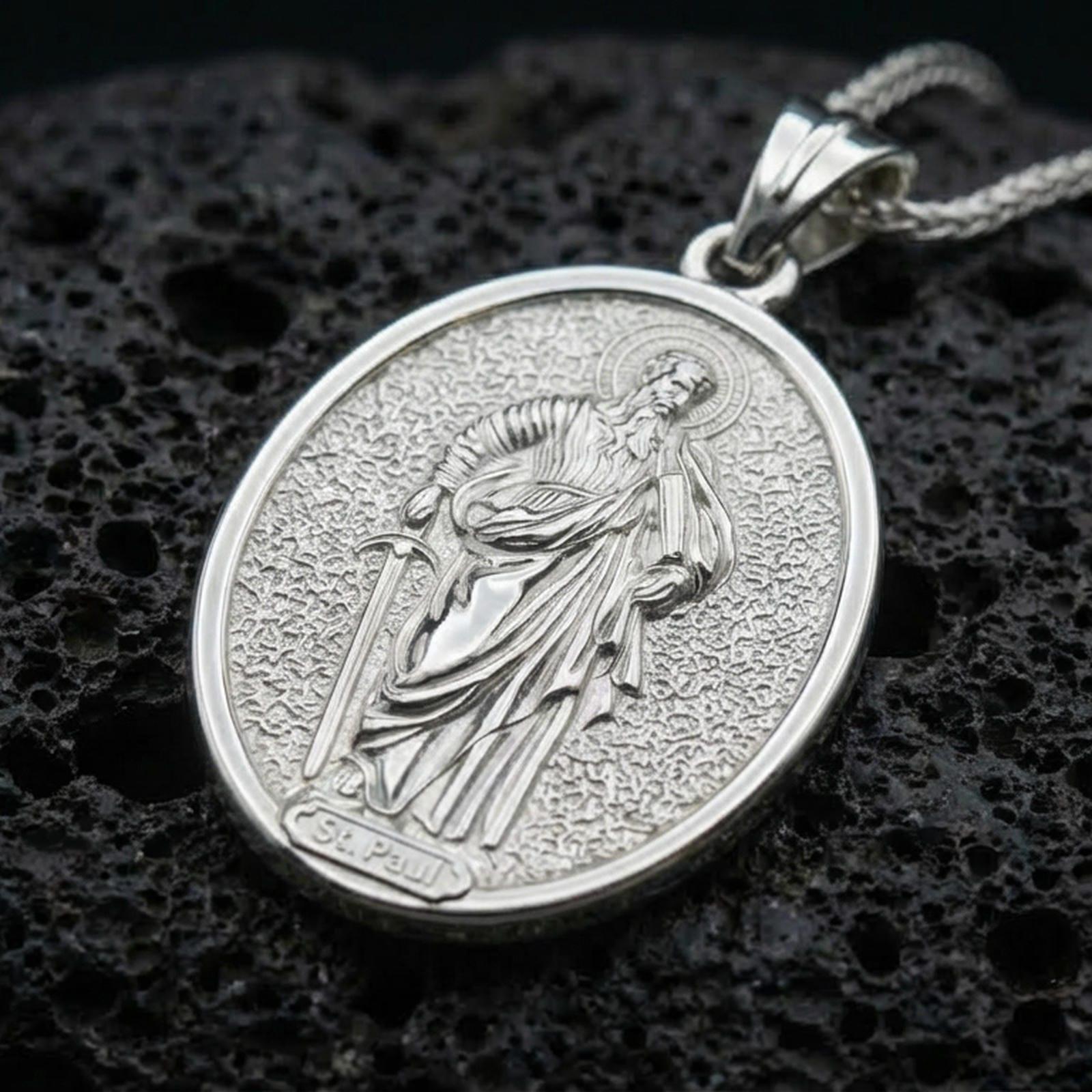 Polished silver Saint Paul medallion with engraved saint relief, halo motif, and detailed oval frame placed on textured volcanic rock in a premium studio product shot
