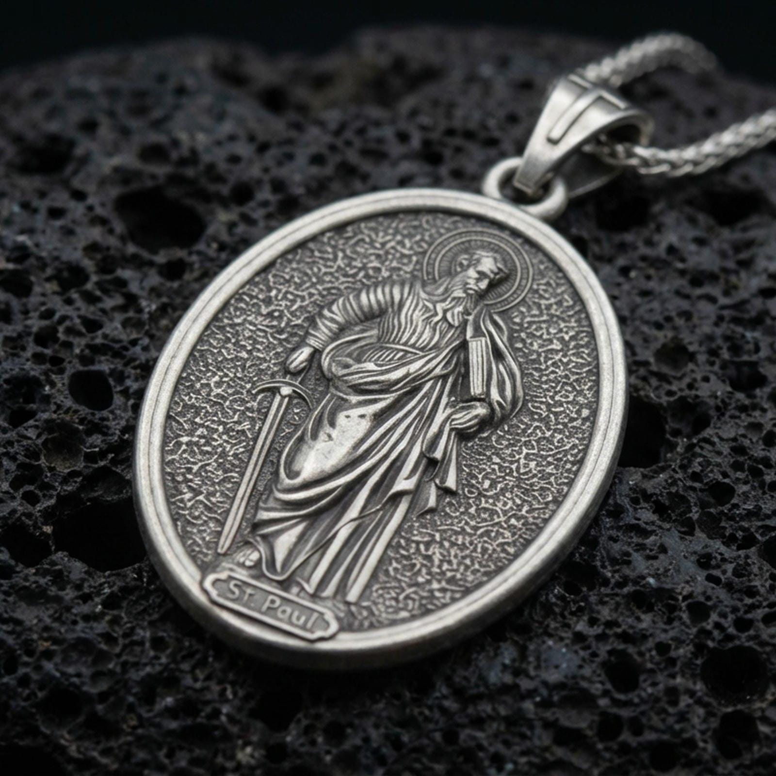 Oxidized Saint Paul pendant with haloed saint figure, sword detail, and oval devotional portrait displayed on dark volcanic rock in a close-up luxury jewelry composition