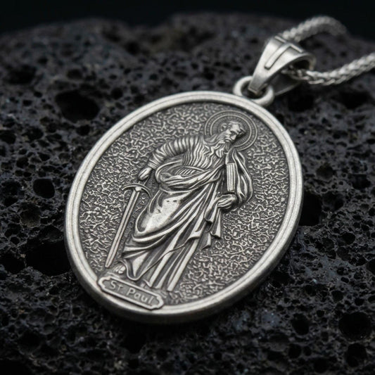 Oxidized Saint Paul pendant with haloed saint figure, sword detail, and oval devotional portrait displayed on dark volcanic rock in a close-up luxury jewelry composition