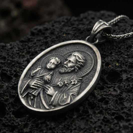 Oxidized Saint Joseph pendant with Child Jesus, halo details, and oval devotional portrait displayed on dark volcanic rock in a close-up luxury jewelry composition