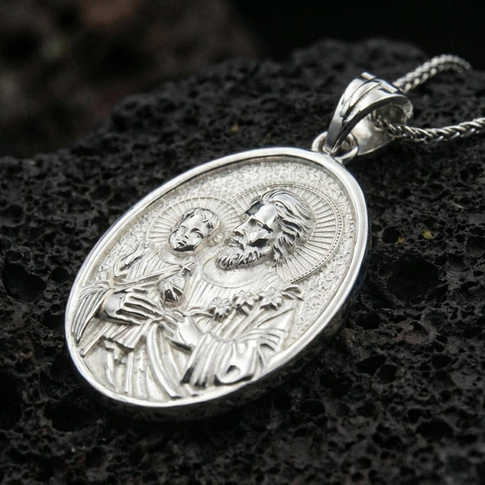 Polished silver Saint Joseph medallion with Child Jesus, sacred halo relief, and detailed oval frame placed on textured volcanic rock in a premium studio product shot