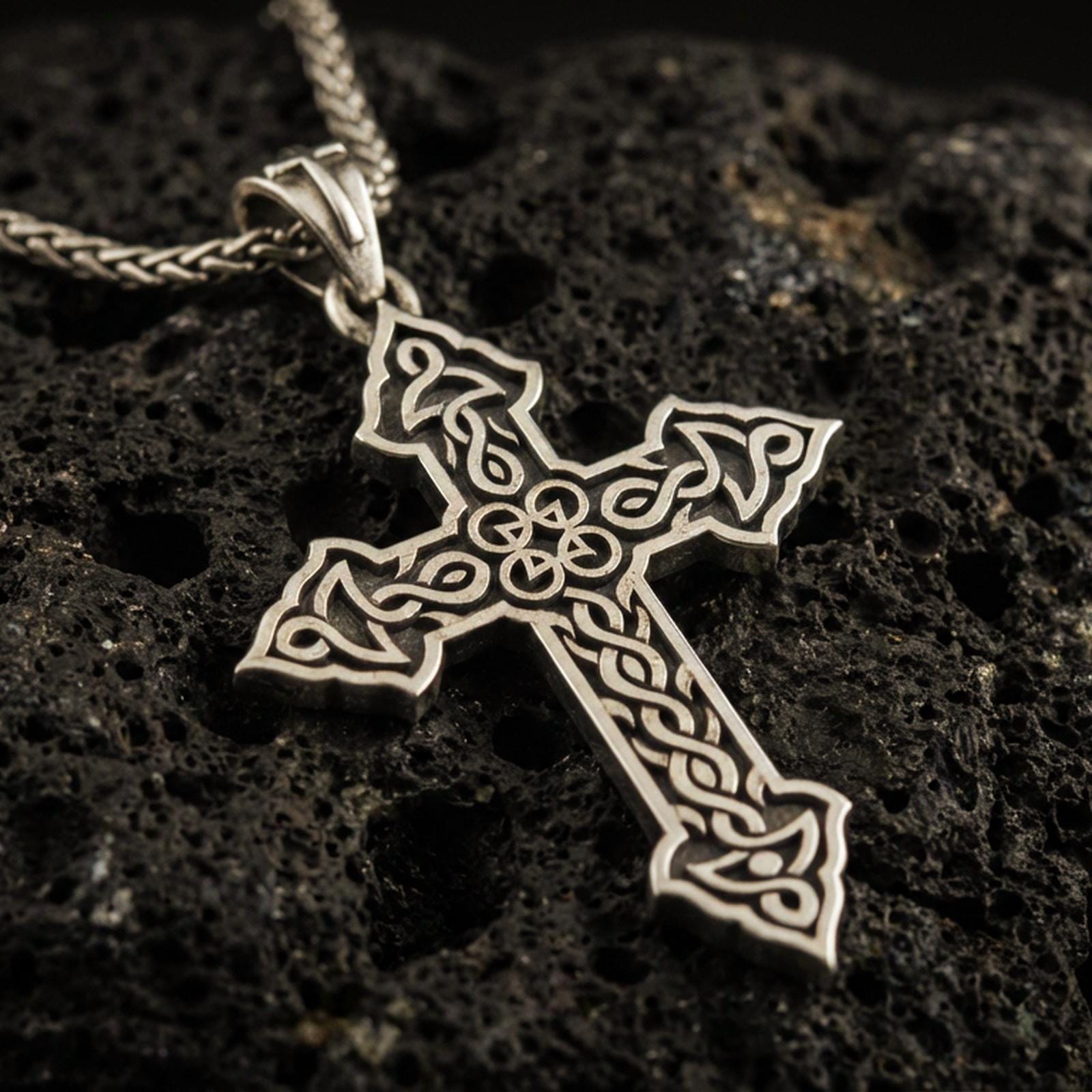 Bronze Celtic cross pendant with intricate knotwork details and open ornamental silhouette displayed on dark volcanic rock in a close-up luxury jewelry composition
