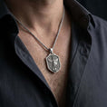 Load image into Gallery viewer, Oxidized Saint Michael shield pendant necklace worn on the neck, styled in a dark lifestyle portrait with an open shirt and masculine presentation.
