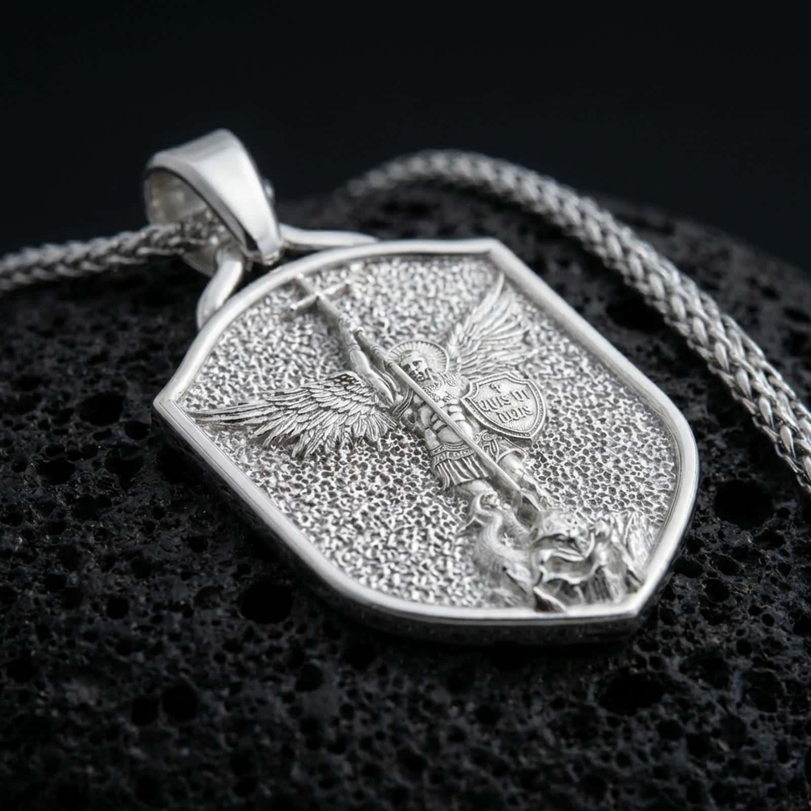 Polished silver Saint Michael shield pendant necklace displayed on black volcanic rock, shown in a refined macro product shot with detailed archangel relief and protective shield motif.