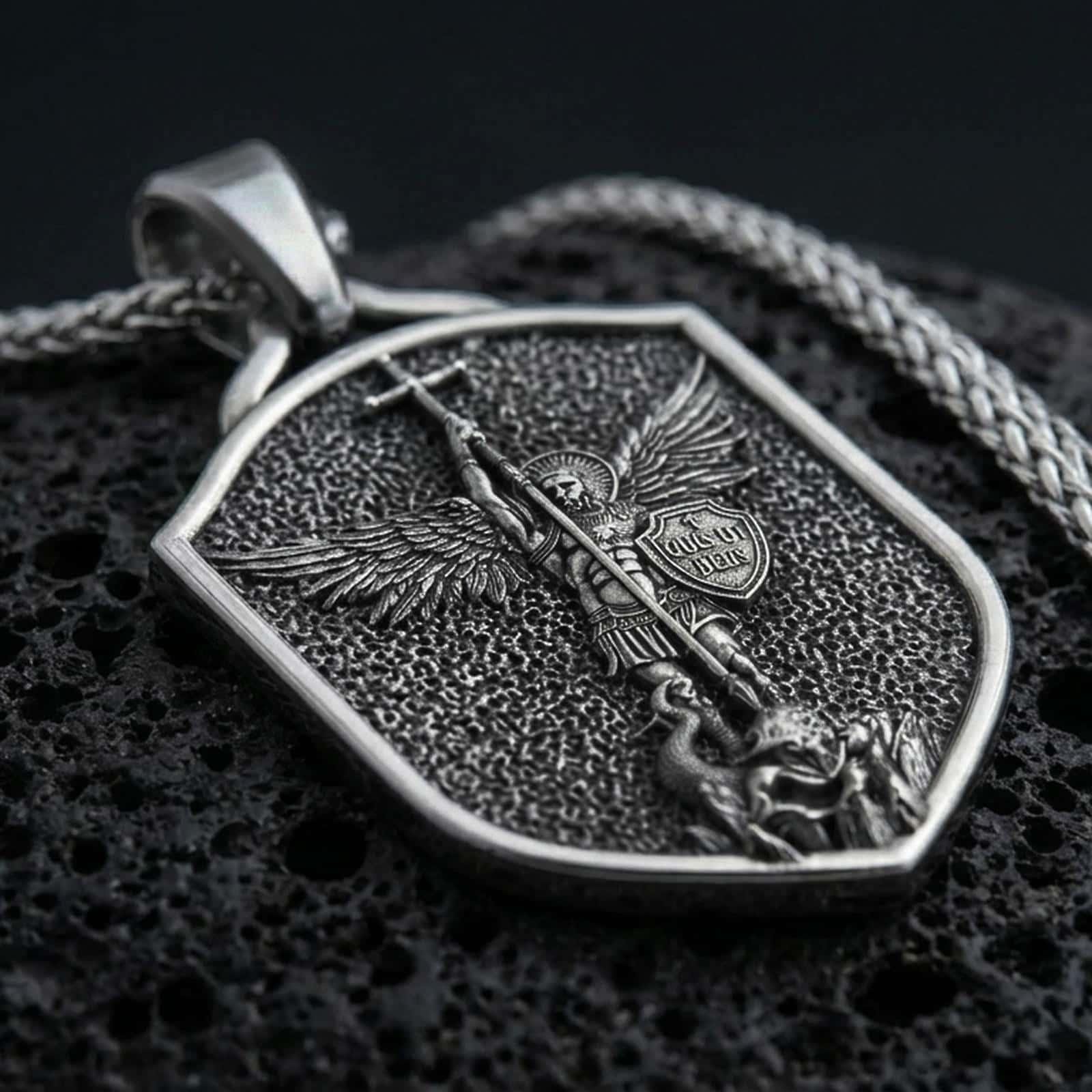 Sterling silver Saint Michael shield pendant necklace with an oxidized finish resting on textured volcanic rock, captured in a close-up premium studio composition.