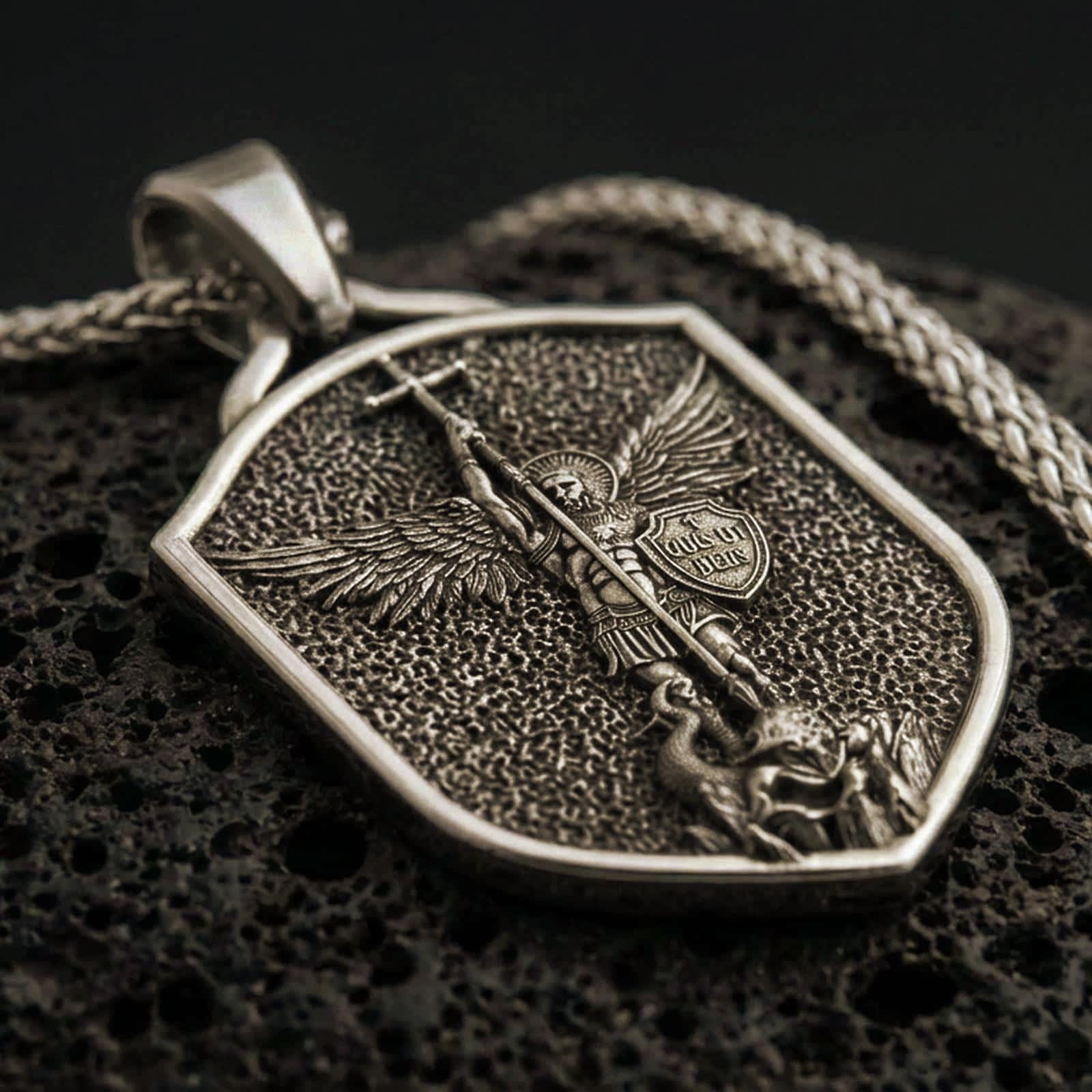 Bronze Saint Michael shield pendant necklace with an oxidized finish resting on textured volcanic rock, captured in a close-up premium studio composition.