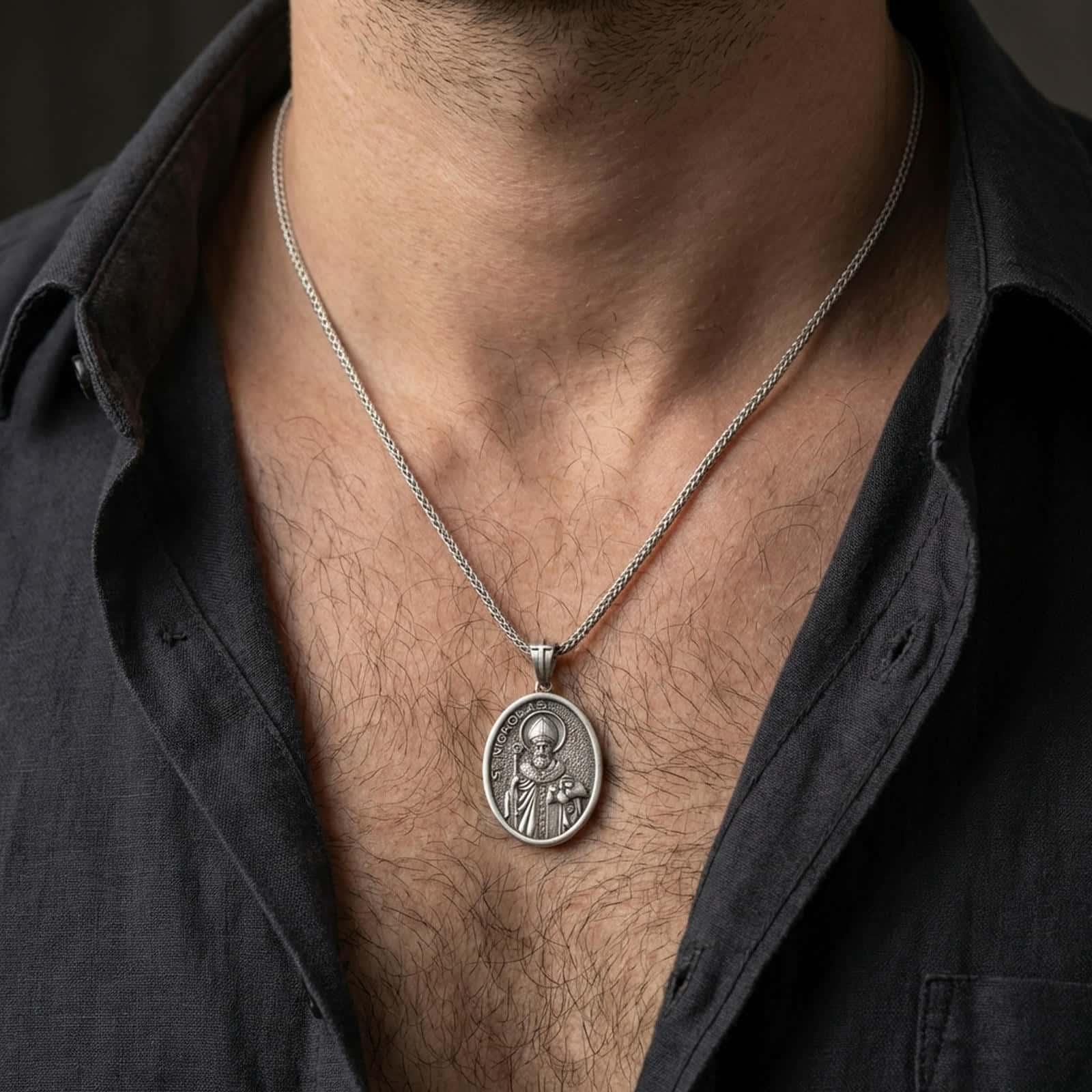 Oxidized Saint Nicholas pendant necklace worn on the neck, styled in a dark lifestyle portrait with an open shirt and masculine presentation.
