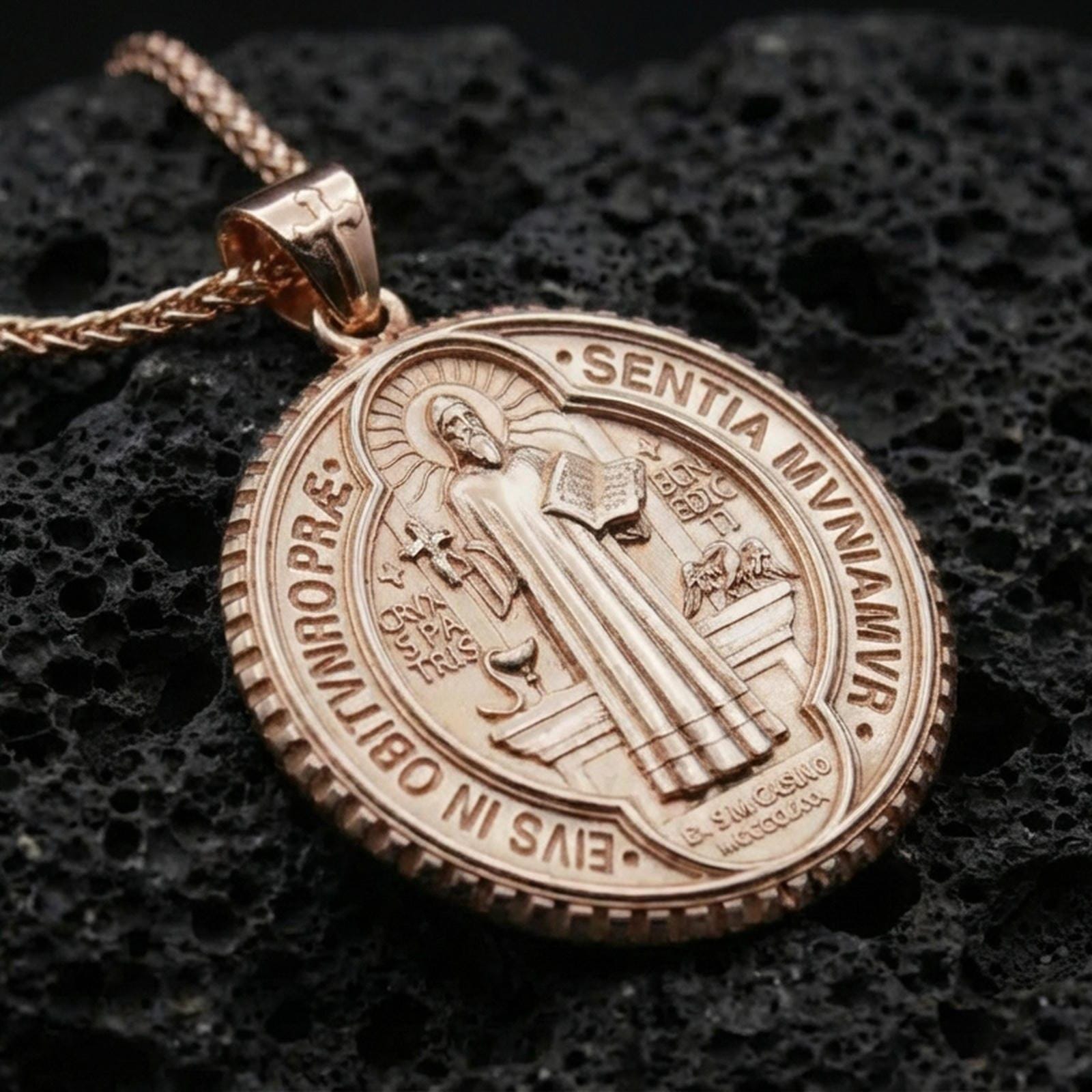 Rose gold plated Saint Benedict necklace with detailed religious iconography, circular medal silhouette, and engraved devotional elements resting on volcanic rock in an elegant close-up product presentation