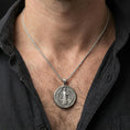 Load image into Gallery viewer, Oxidized Saint Benedict medallion pendant with saint figure and Latin inscriptions worn on the neck in a lifestyle jewelry shot, styled against the chest for a devotional and timeless look
