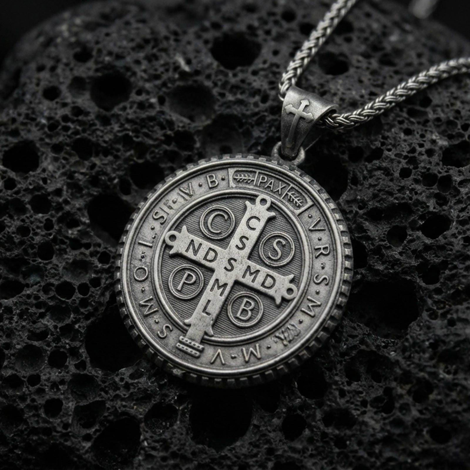 Oxidized Saint Benedict medal pendant with central cross, engraved initials, and round protective medallion design displayed on dark volcanic rock in a close-up luxury jewelry composition