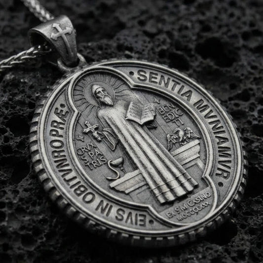 Oxidized Saint Benedict medallion pendant with robed saint figure, Latin inscriptions, and round devotional design displayed on dark volcanic rock in a close-up luxury jewelry composition