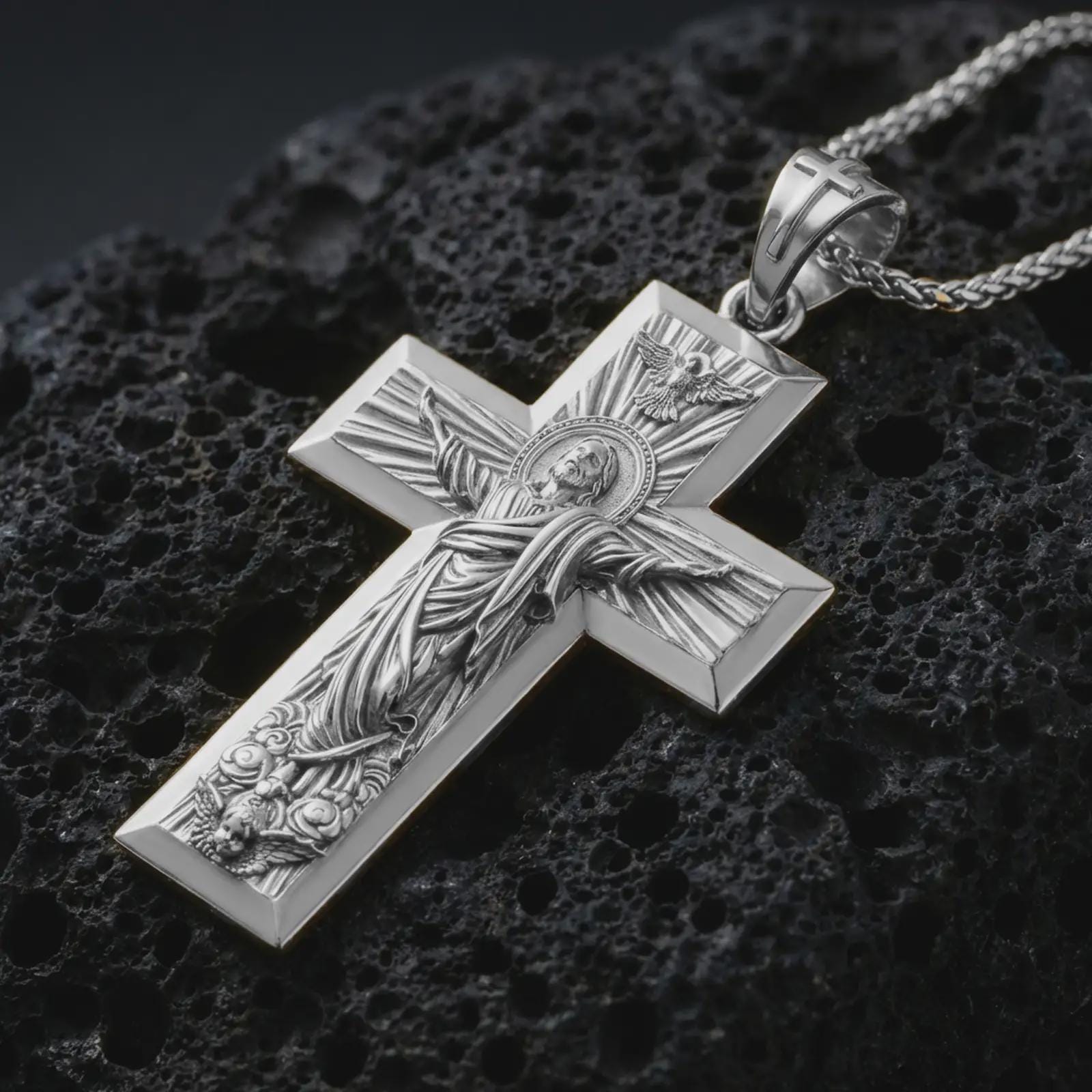Polished silver Jesus Ascension cross pendant necklace displayed on black volcanic rock, shown in a refined macro product shot with detailed sacred figure and radiant cross relief.