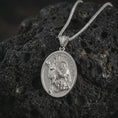 Load image into Gallery viewer, Polished silver Saint Hubert pendant necklace displayed on black volcanic rock, shown in a refined macro product shot with detailed hunter saint and stag relief.
