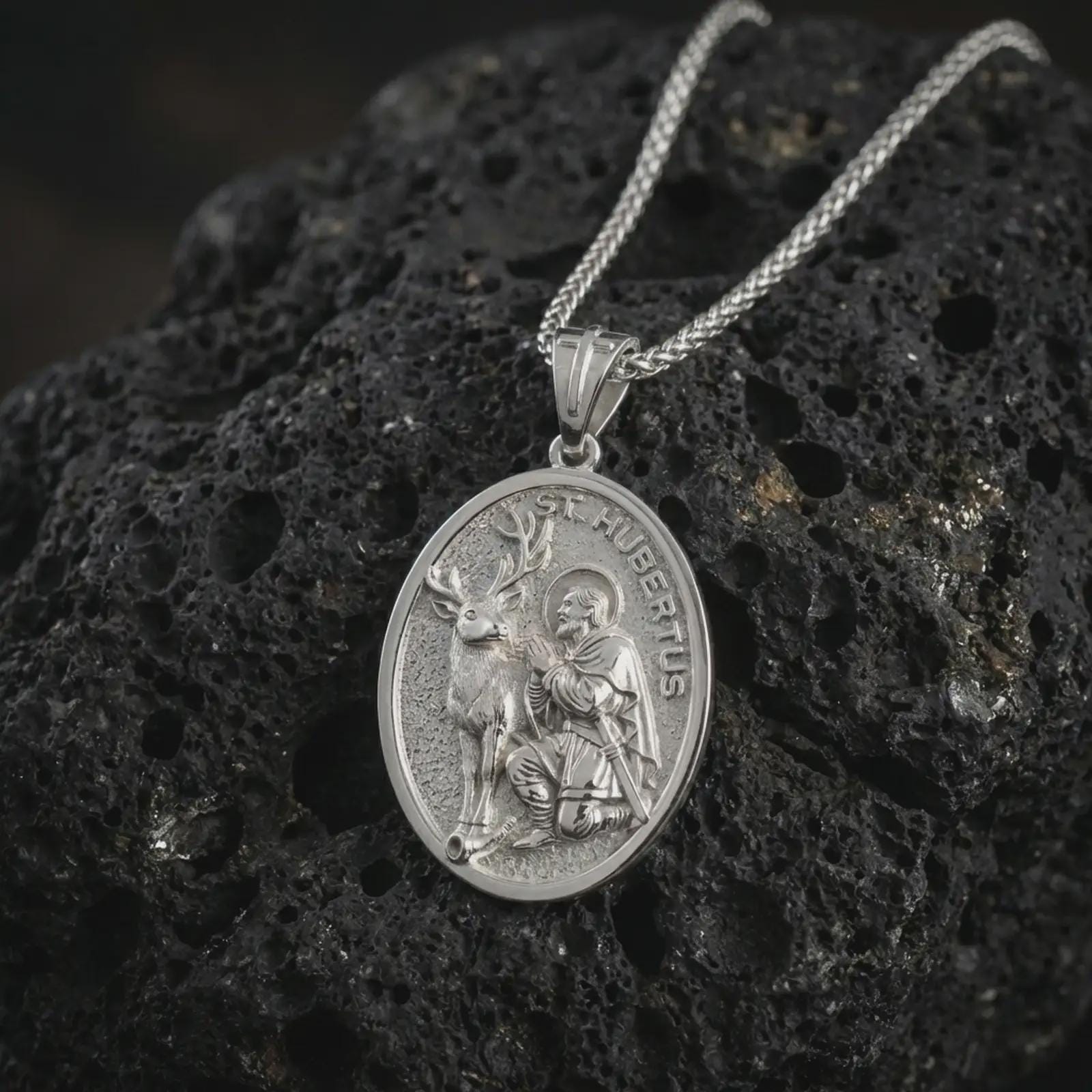 Polished silver Saint Hubert pendant necklace displayed on black volcanic rock, shown in a refined macro product shot with detailed hunter saint and stag relief.