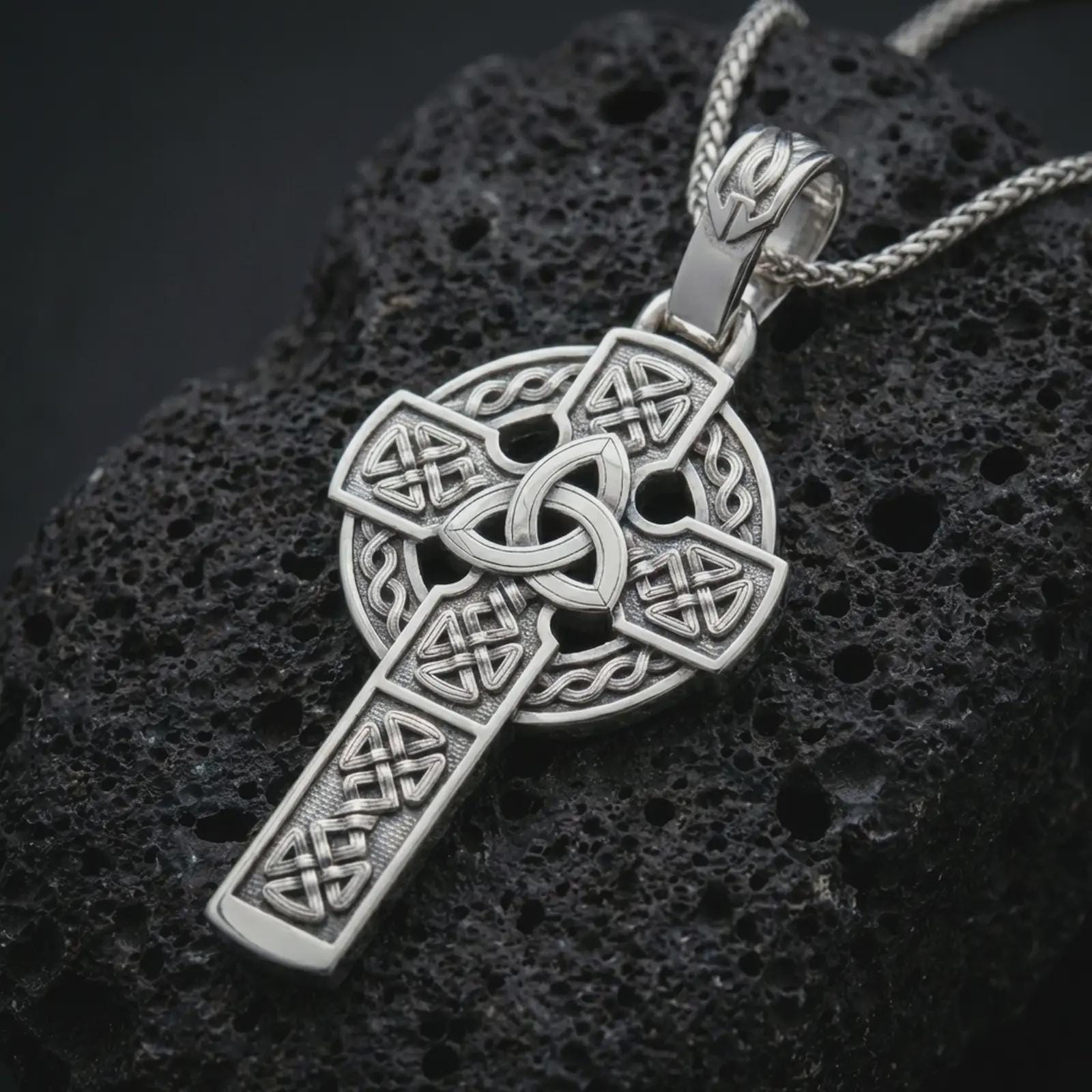 Polished silver Celtic cross pendant necklace displayed on black volcanic rock, shown in a refined macro product shot with Trinity knot and circular halo detail.