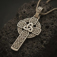 Load image into Gallery viewer, Bronze Celtic cross pendant necklace with an oxidized finish resting on textured volcanic rock, captured in a close-up premium studio composition.
