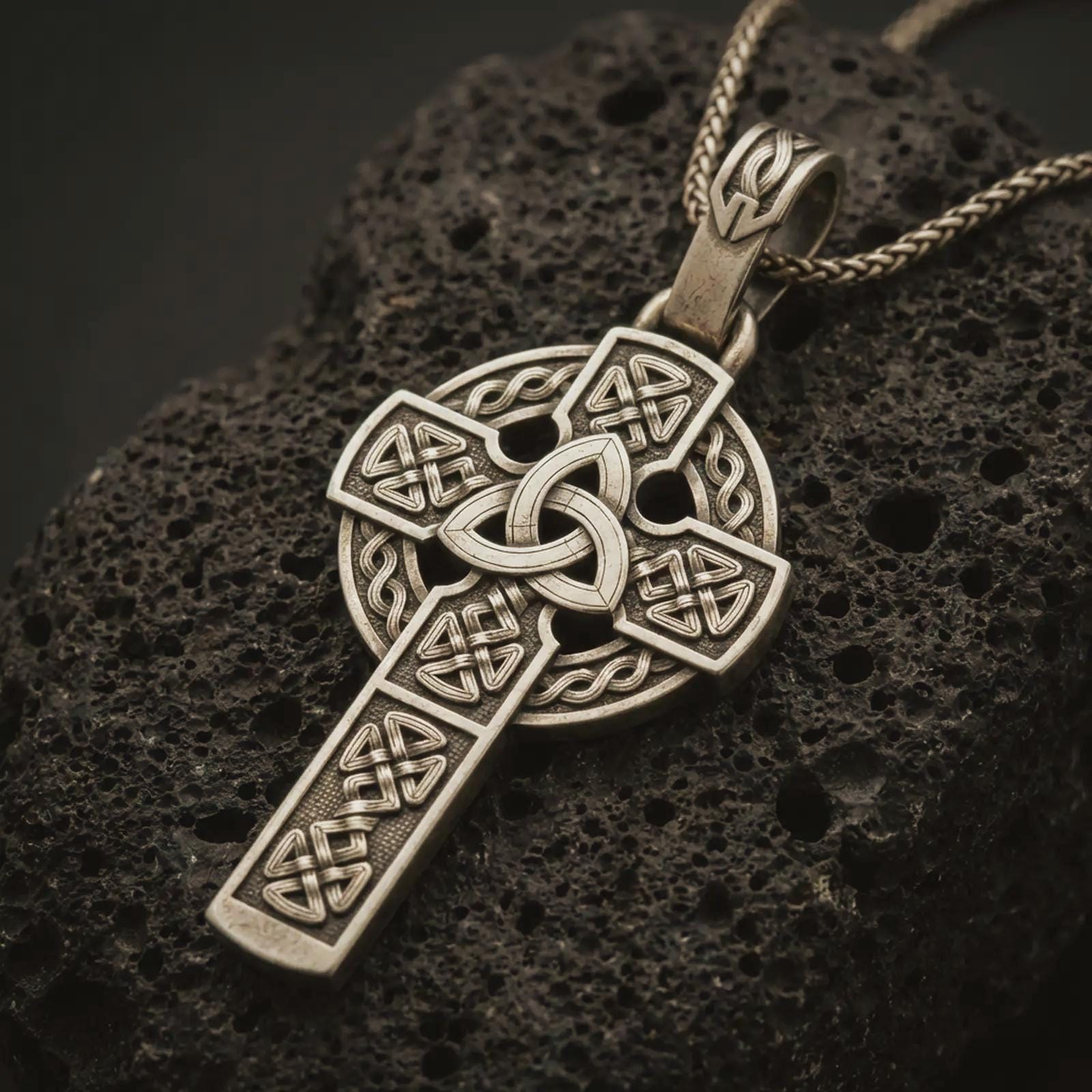 Bronze Celtic cross pendant necklace with an oxidized finish resting on textured volcanic rock, captured in a close-up premium studio composition.