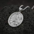 Load image into Gallery viewer, Polished silver Saint Benedict pendant necklace displayed on black volcanic rock, shown in a refined macro product shot with detailed saint figure, staff, and engraved lettering.
