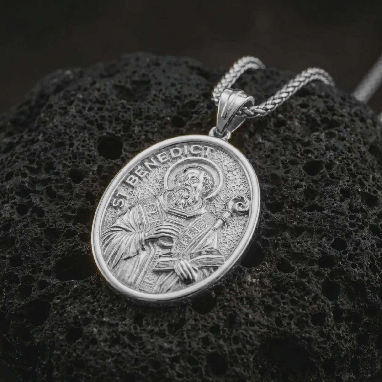 Polished silver Saint Benedict pendant necklace displayed on black volcanic rock, shown in a refined macro product shot with detailed saint figure, staff, and engraved lettering.