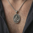 Load image into Gallery viewer, Oxidized Saint Benedict pendant necklace worn on the neck, styled in a dark lifestyle portrait with an open shirt and masculine presentation.
