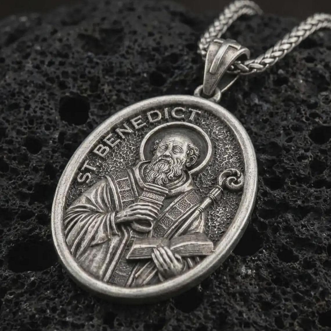 Sterling silver Saint Benedict pendant necklace with an oxidized finish resting on textured volcanic rock, captured in a close-up premium studio composition.