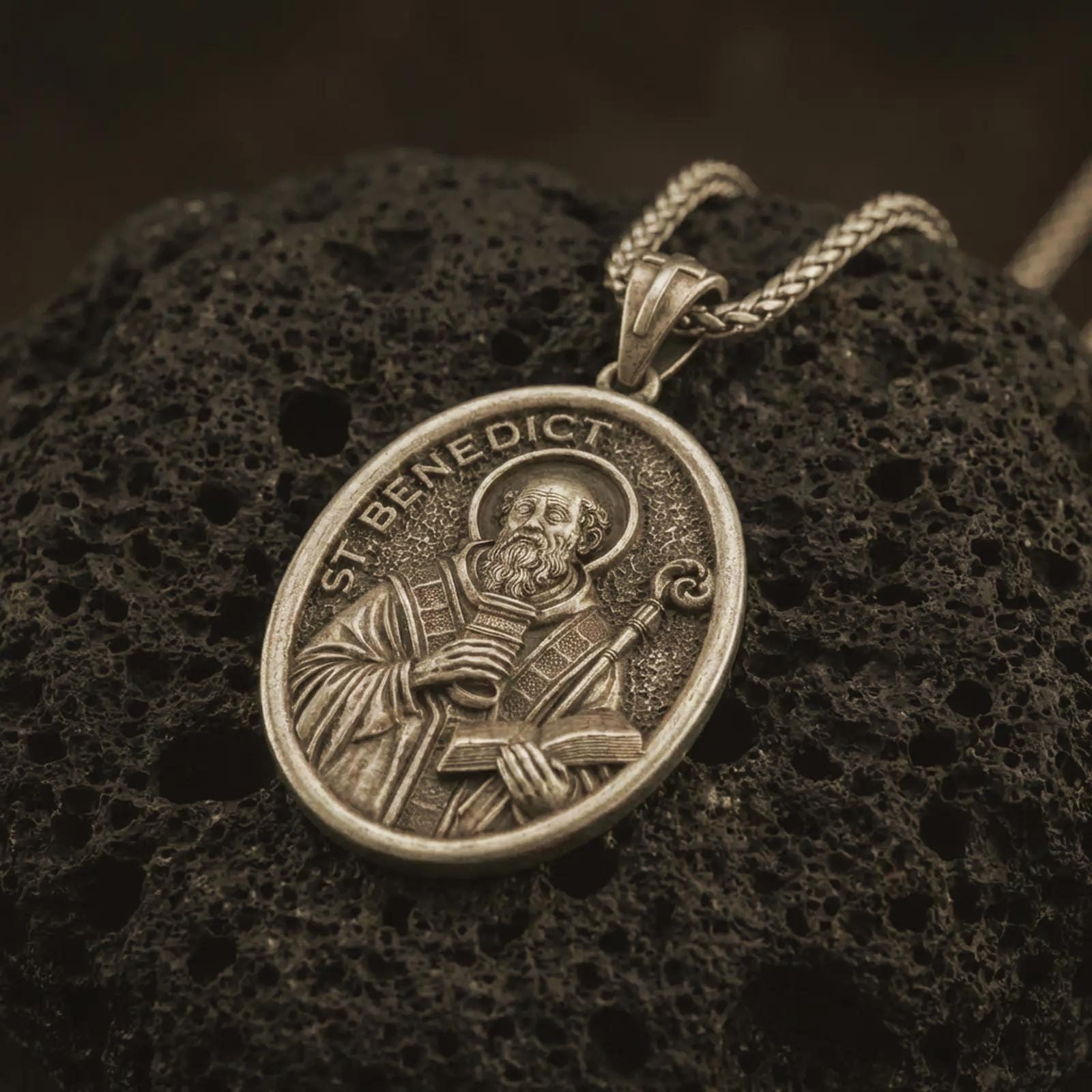 Bronze Saint Benedict pendant necklace with an oxidized finish resting on textured volcanic rock, captured in a close-up premium studio composition.
