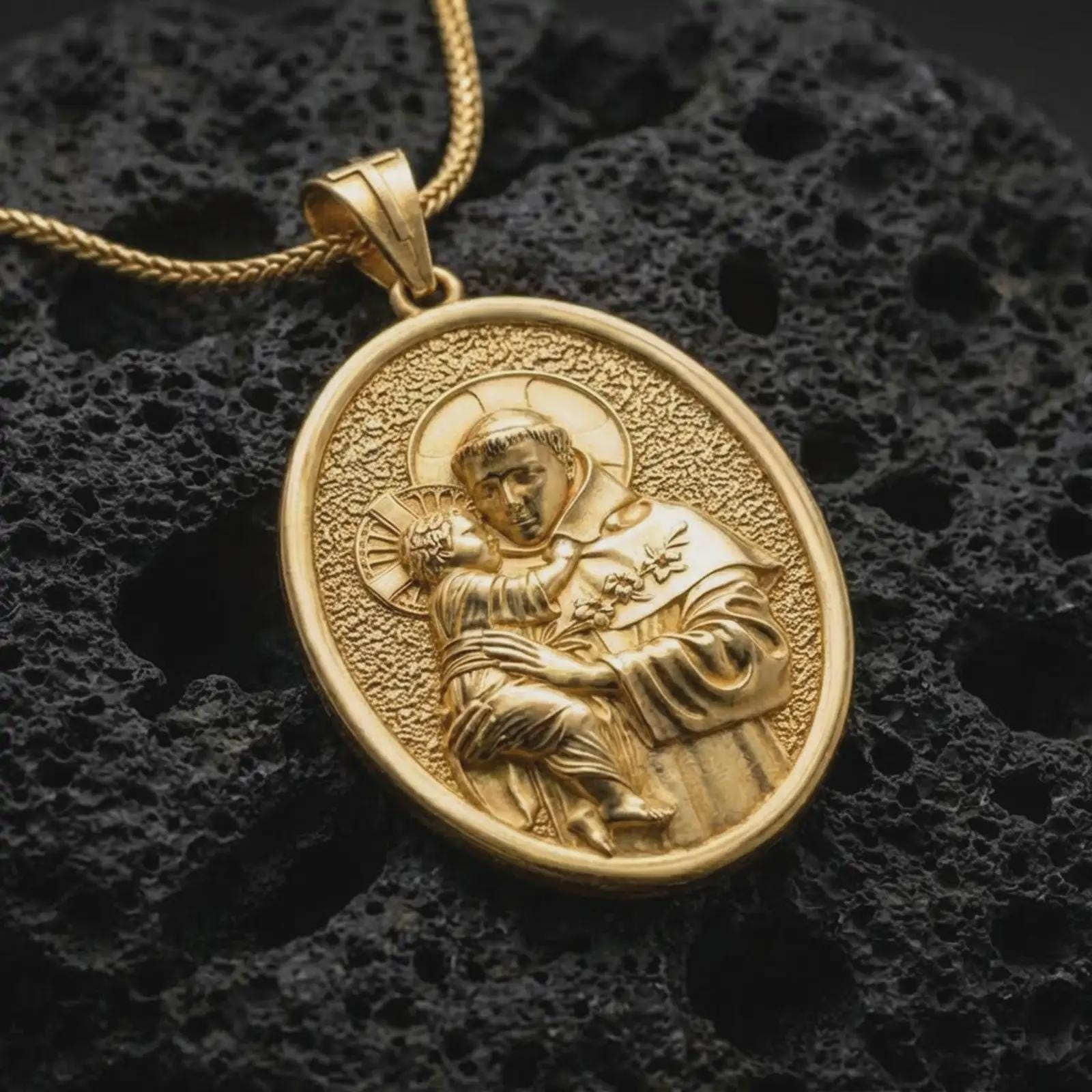 Gold plated Saint Anthony pendant necklace presented on porous volcanic rock, highlighting the oval medallion form and sacred devotional design in a luxurious close-up jewelry image.