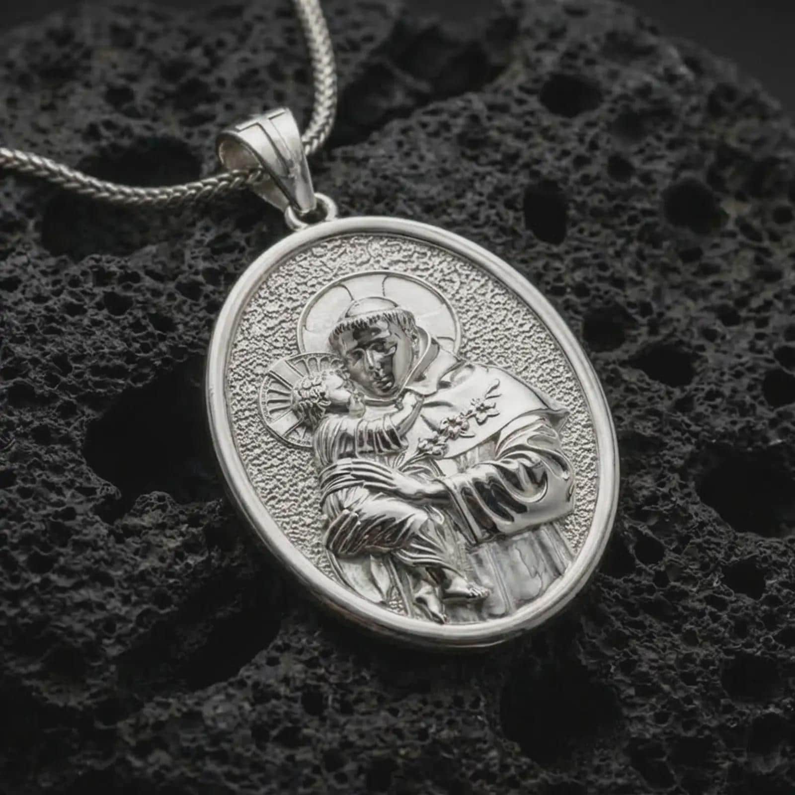 Polished silver Saint Anthony pendant necklace displayed on black volcanic rock, shown in a refined macro product shot with detailed saint and child relief.