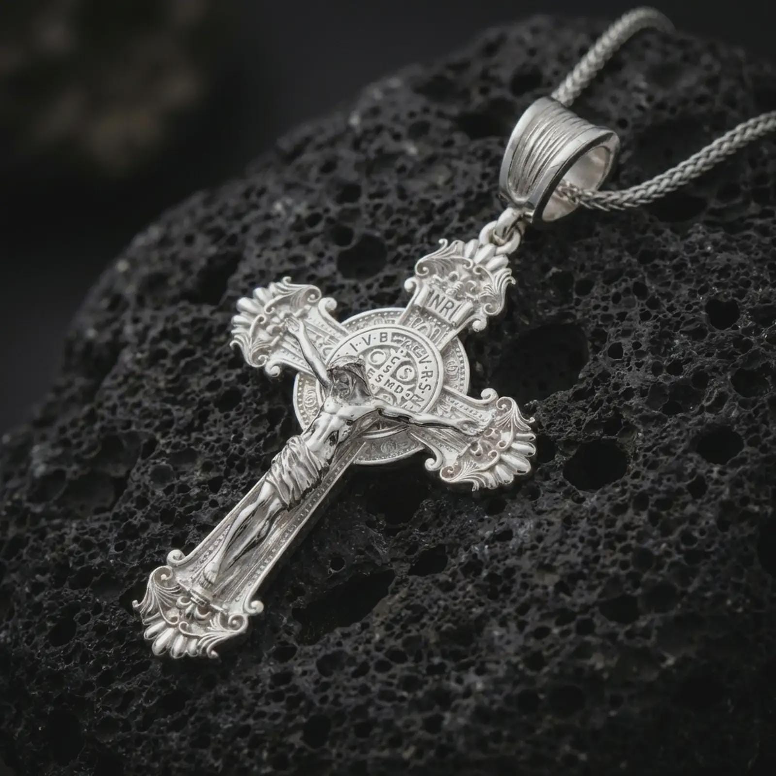 Polished silver Saint Benedict crucifix necklace displayed on black volcanic rock, shown in a refined macro product shot with detailed corpus figure and medallion center.