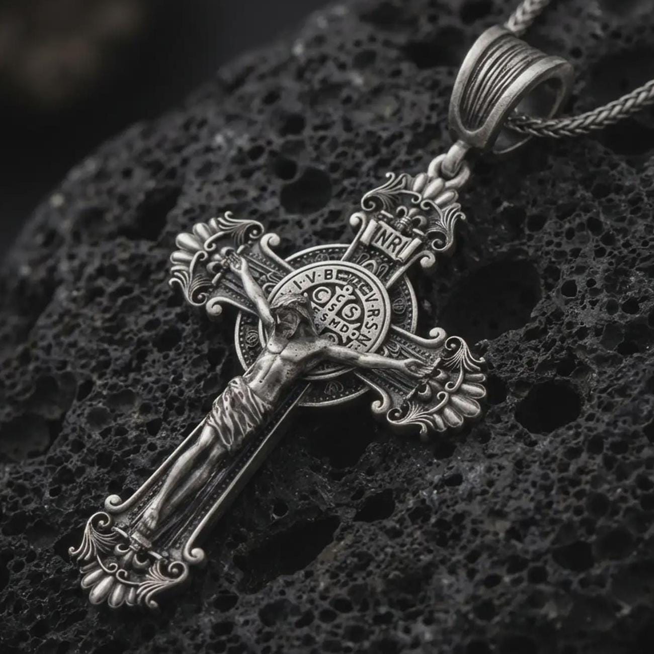 Sterling silver Saint Benedict crucifix necklace with an oxidized finish resting on textured volcanic rock, captured in a close-up premium studio composition.