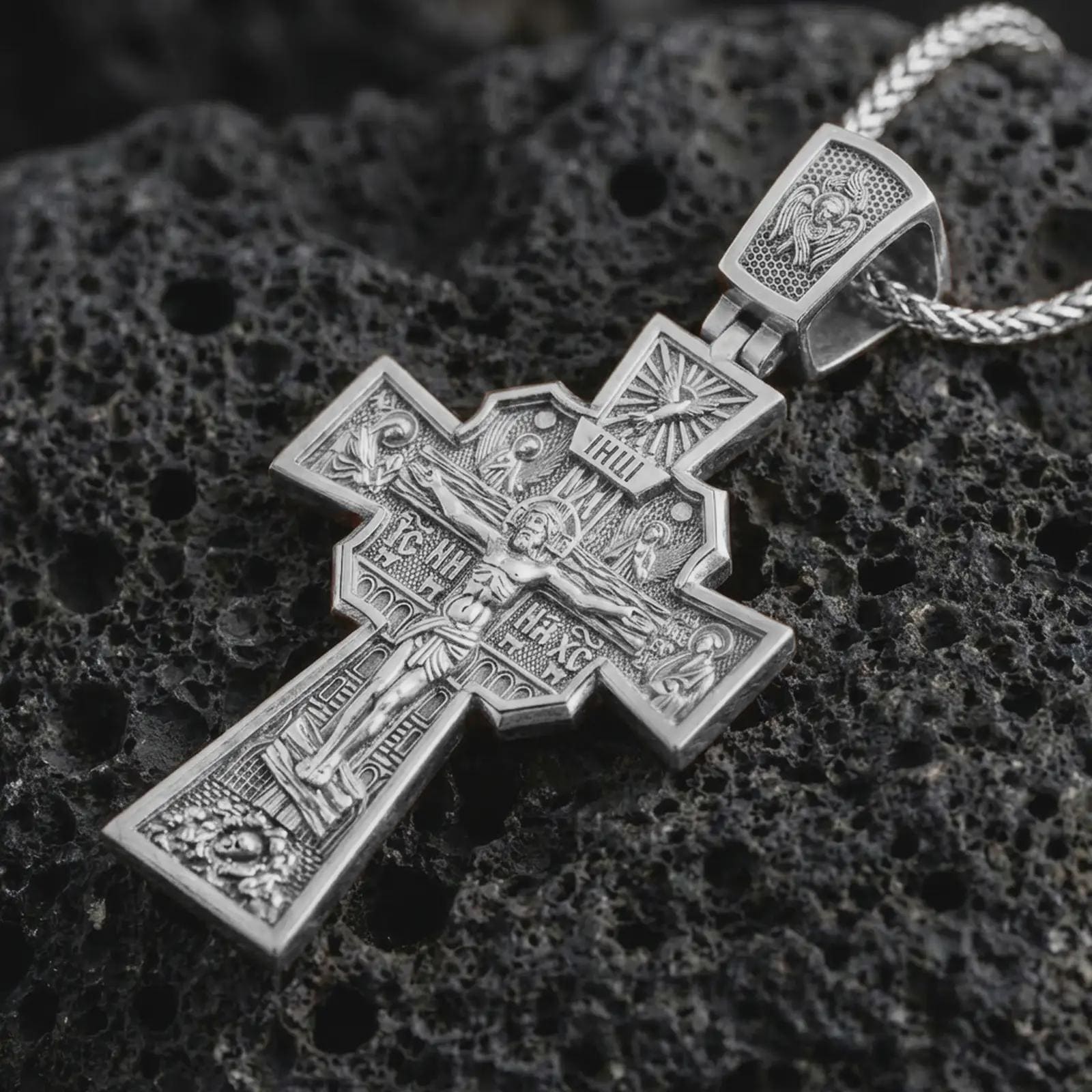 Polished silver Orthodox cross pendant necklace displayed on black volcanic rock, shown in a refined macro product shot with detailed crucifix figure and sacred engraved iconography.