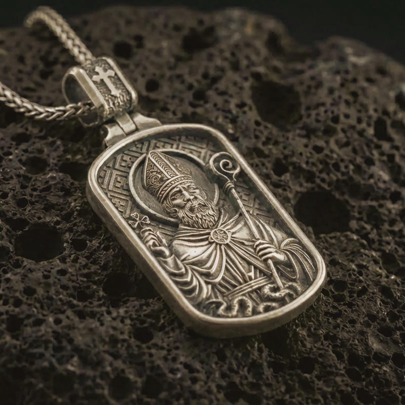 Bronze Saint Patrick dog tag pendant necklace with an oxidized finish resting on textured volcanic rock, captured in a close-up premium studio composition.