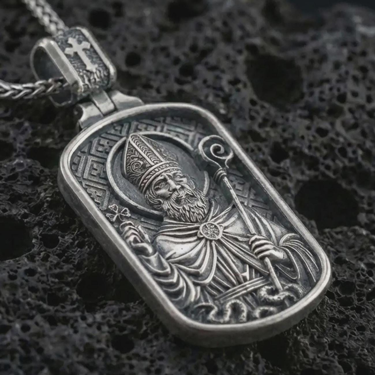 Sterling silver Saint Patrick dog tag pendant necklace with an oxidized finish resting on textured volcanic rock, captured in a close-up premium studio composition.