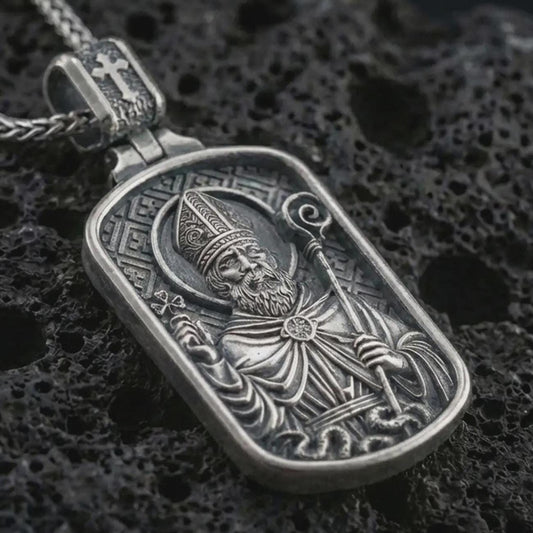 Sterling silver Saint Patrick dog tag pendant necklace with an oxidized finish resting on textured volcanic rock, captured in a close-up premium studio composition.