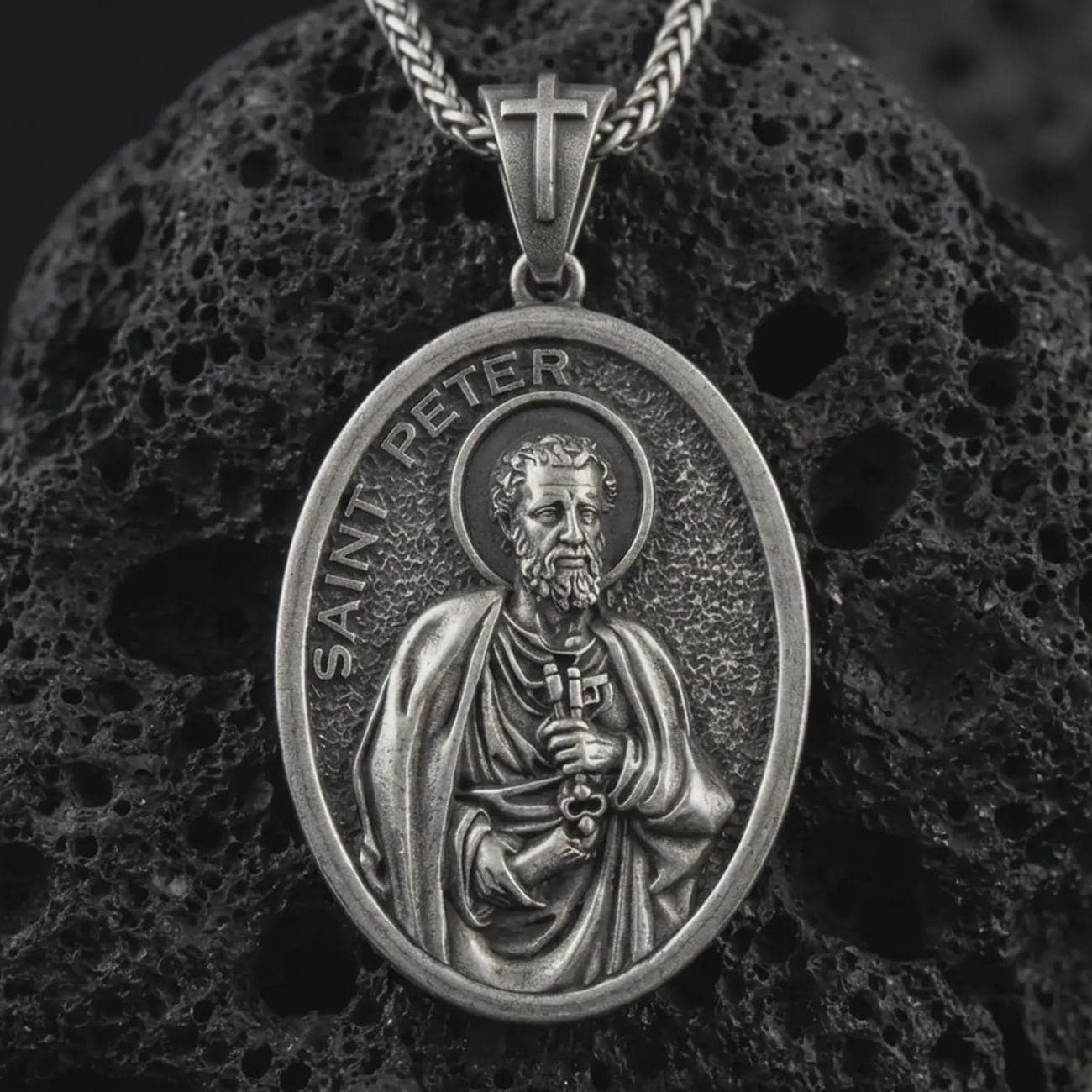 Sterling silver Saint Peter pendant necklace with an oxidized finish resting on textured volcanic rock, captured in a close-up premium studio composition.