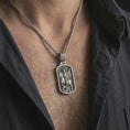 Load image into Gallery viewer, Oxidized Saint George dog tag pendant necklace worn on the neck, styled in a dark lifestyle portrait with an open shirt and masculine presentation.
