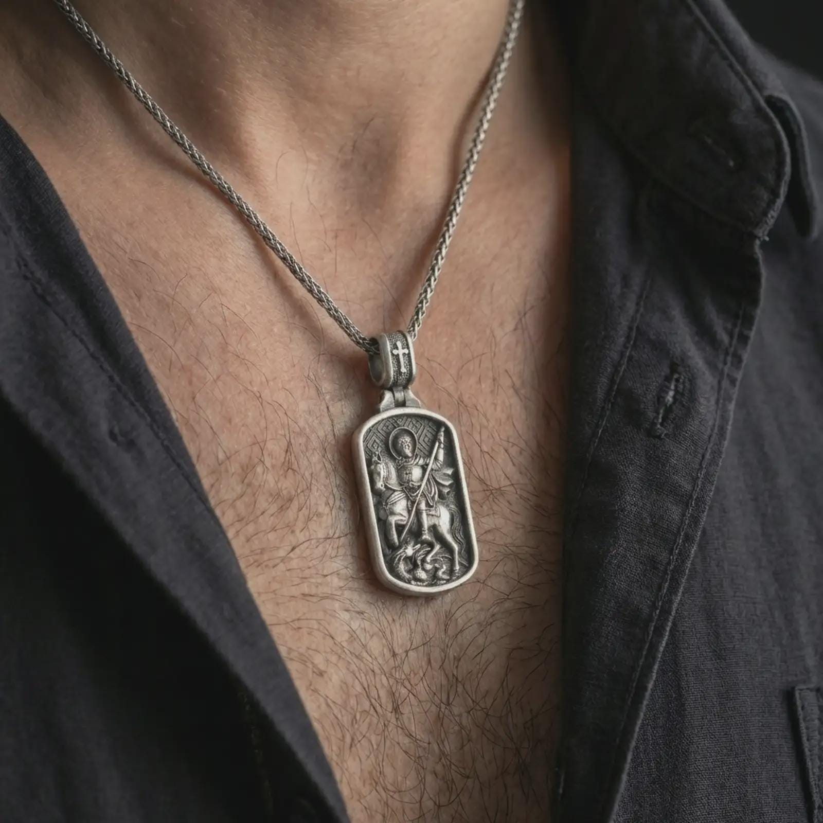 Oxidized Saint George dog tag pendant necklace worn on the neck, styled in a dark lifestyle portrait with an open shirt and masculine presentation.