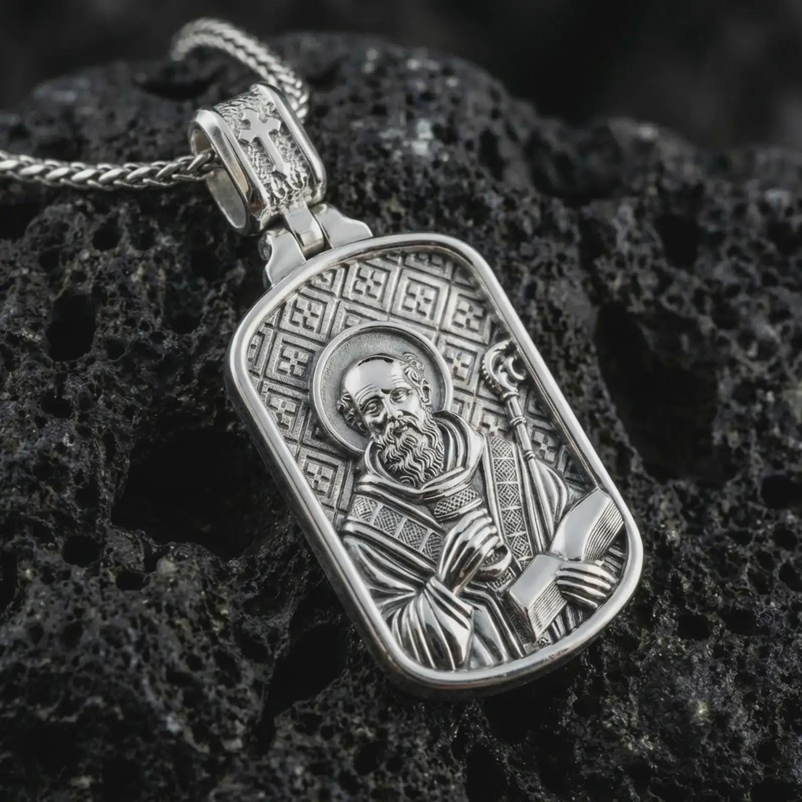 Polished silver Saint Benedict dog tag pendant necklace displayed on black volcanic rock, shown in a refined macro product shot with detailed saint figure, staff, book, and engraved background pattern.
