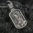 Load image into Gallery viewer, Sterling silver Saint Benedict dog tag pendant necklace with an oxidized finish resting on textured volcanic rock, captured in a close-up premium studio composition.
