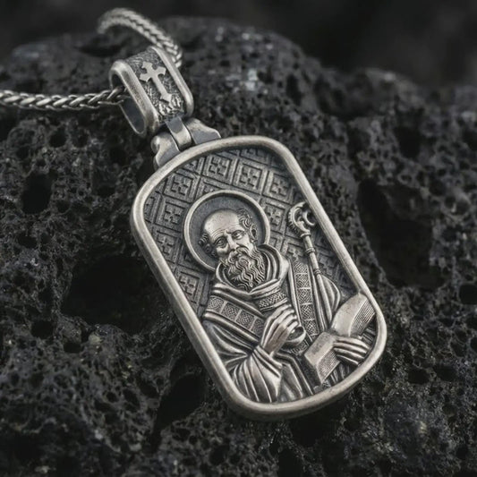 Sterling silver Saint Benedict dog tag pendant necklace with an oxidized finish resting on textured volcanic rock, captured in a close-up premium studio composition.
