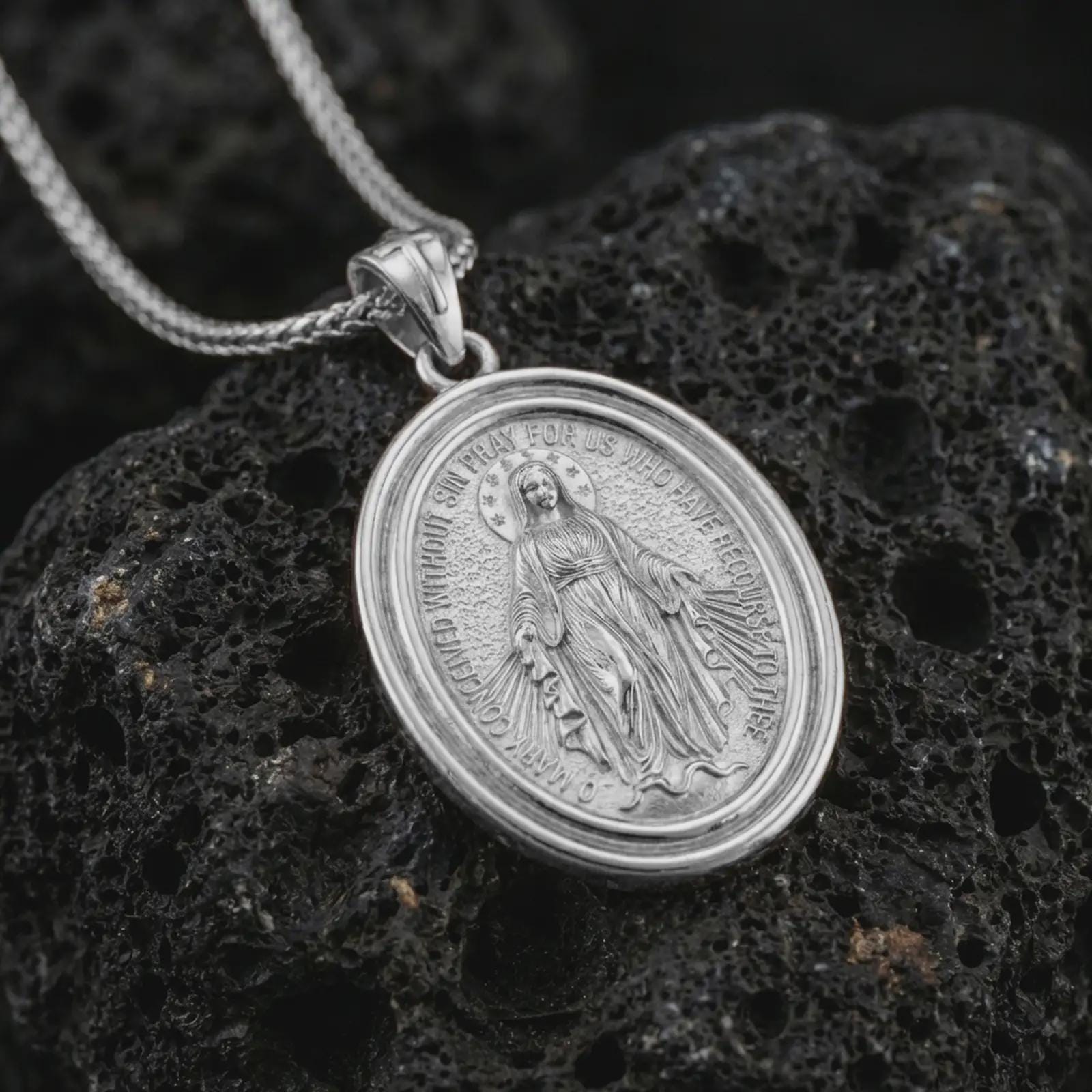 Polished silver Miraculous Medal pendant necklace displayed on black volcanic rock, shown in a refined macro product shot with detailed Virgin Mary figure and engraved prayer lettering.