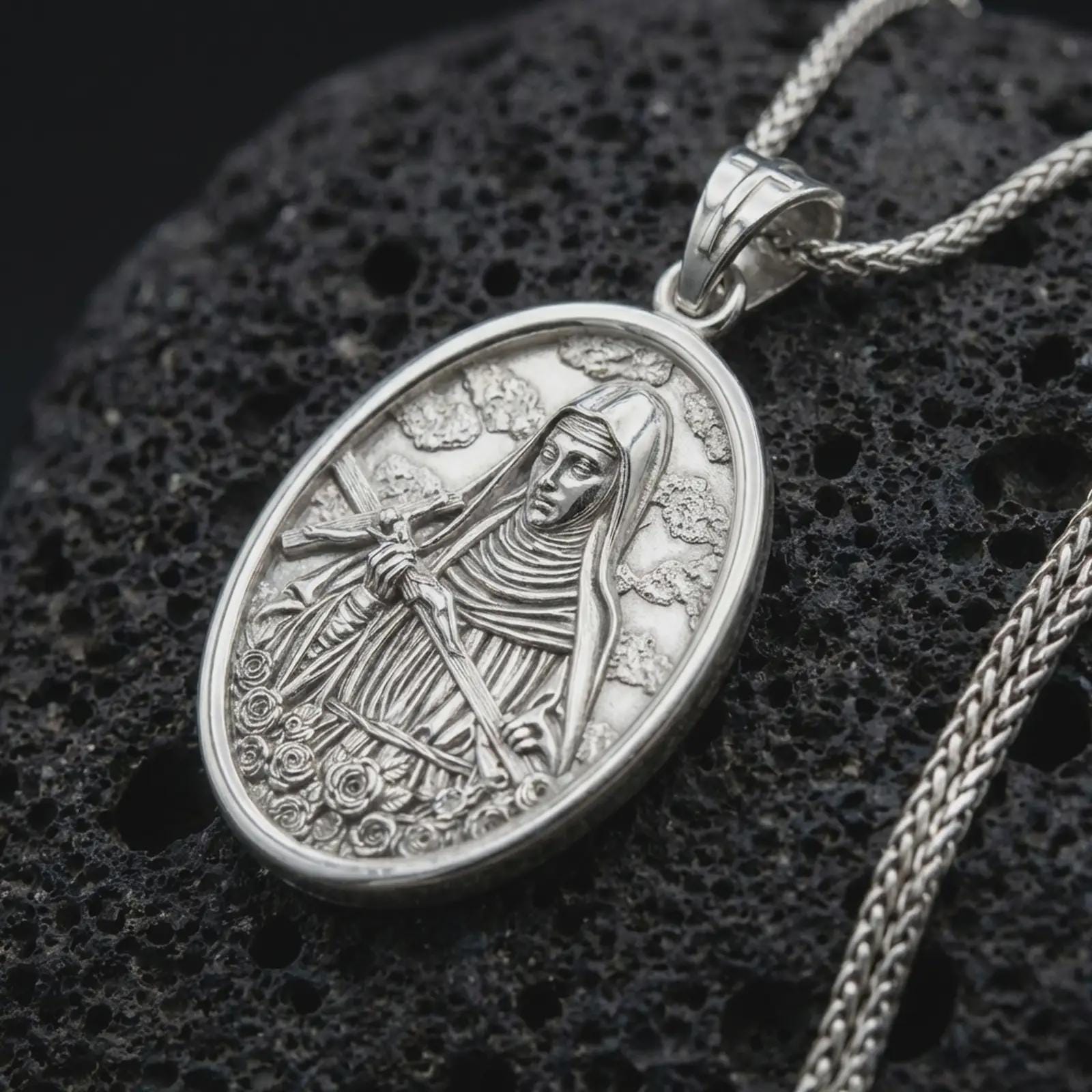 Polished silver Saint Rita pendant necklace displayed on black volcanic rock, shown in a refined macro product shot with detailed saint figure, crucifix, and floral relief.