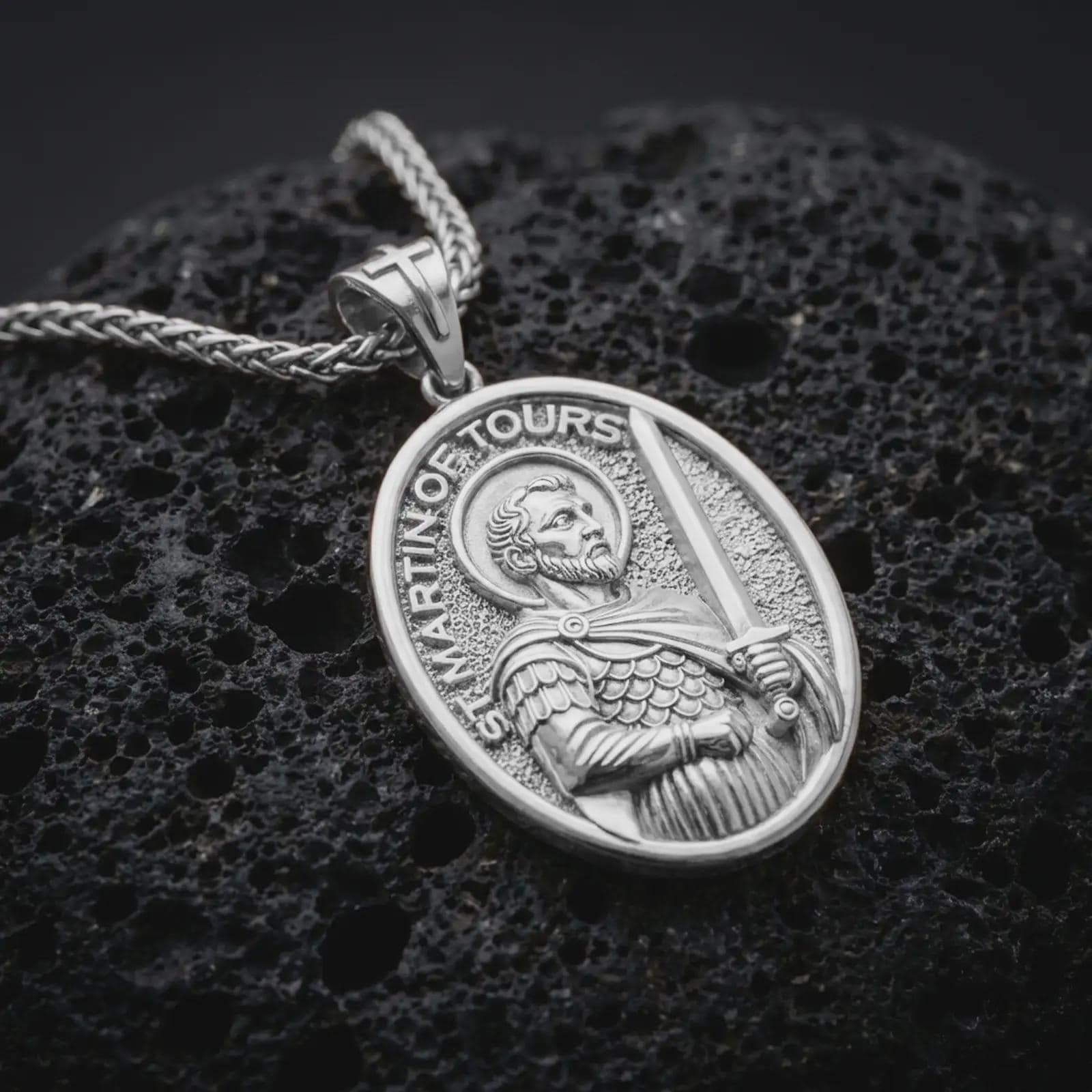 Polished silver Saint Martin of Tours pendant necklace displayed on black volcanic rock, shown in a refined macro product shot with detailed saint figure, sword, and engraved lettering.