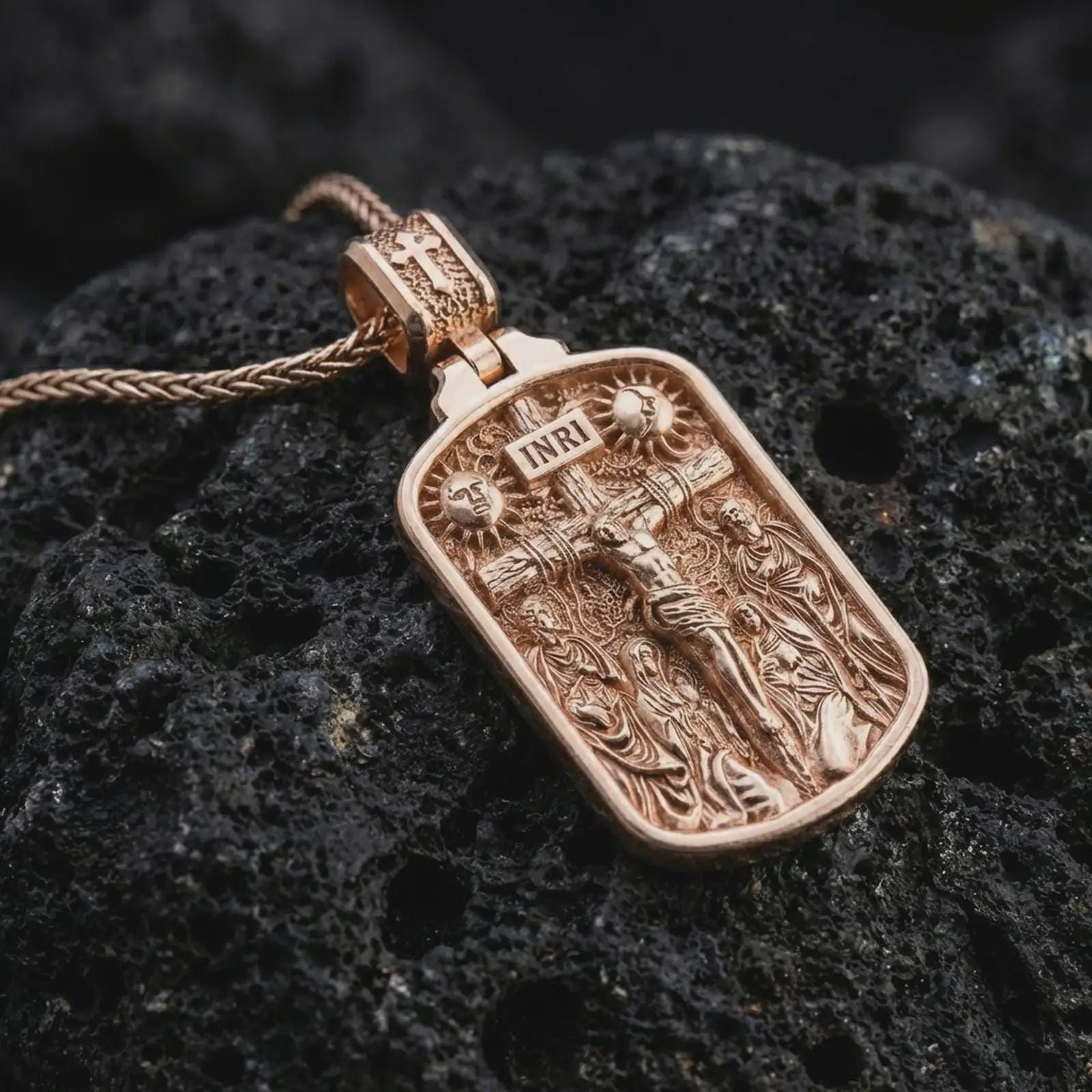 Rose gold plated Jesus INRI crucifixion pendant necklace on dark volcanic rock, photographed in a detailed premium studio macro view.
