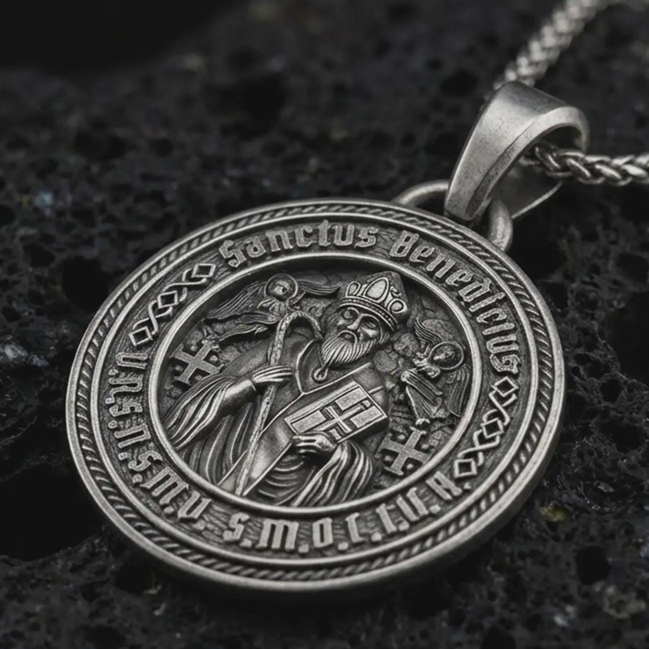 Sterling silver Saint Benedict medal pendant necklace with an oxidized finish resting on textured volcanic rock, captured in a close-up premium studio composition.