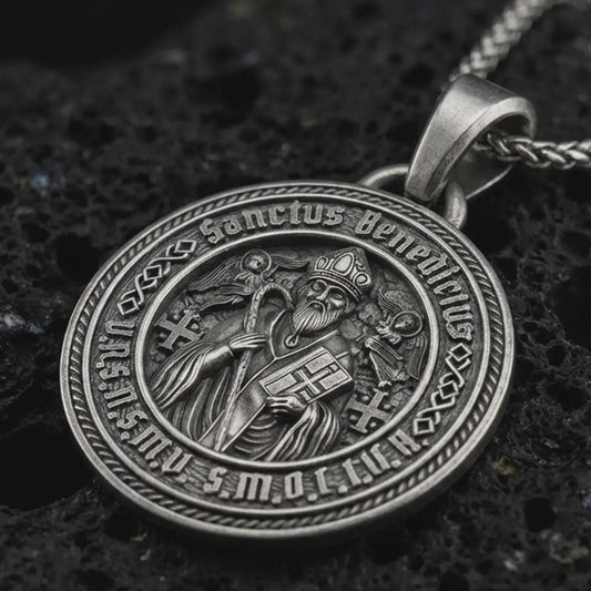 Sterling silver Saint Benedict medal pendant necklace with an oxidized finish resting on textured volcanic rock, captured in a close-up premium studio composition.