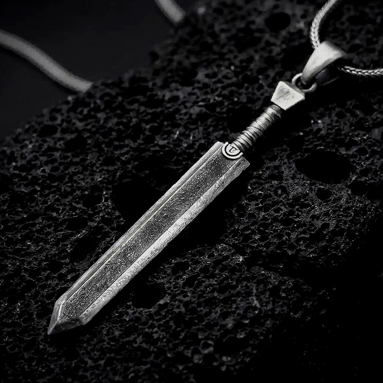 Sterling silver Dragonslayer sword pendant necklace with an oxidized finish resting on textured volcanic rock, captured in a close-up premium studio composition.
